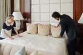 Young head manager of hotel helping chambermaid prepare room for new guests while both bending over double bed with cushions